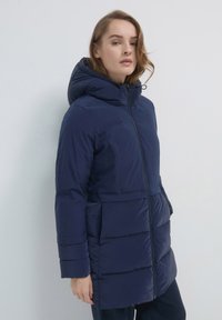 Donna che indossa un cappotto invernale imbottito blu navy con cappuccio, in piedi contro uno sfondo chiaro e uniforme.