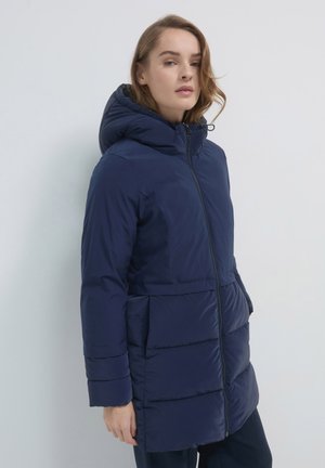 Donna che indossa un cappotto invernale imbottito blu navy con cappuccio, in piedi contro uno sfondo chiaro e uniforme.
