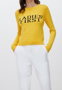 Geel trui met zwarte tekst "LADIES FIRST", geribbelde halslijn en mouwen, aansluitend ontwerp, gecombineerd met witte broek, gladde textuur.