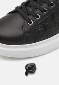 Svarta lädersneakers med texturerat mönster, vit mellansula, silverfärgade öglor och en svart tecknad karaktärs-nål som ligger bredvid.