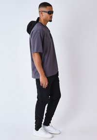 Camiseta corta de manga corta morada con capucha negra, combinada con pantalones cargo negros y zapatillas blancas. Los accesorios incluyen gafas de sol y un collar.
