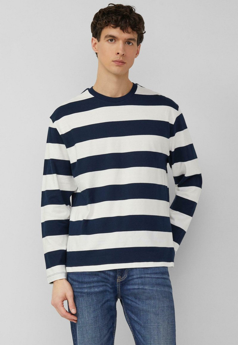 Noor mees lühikeste lokkis juustega, kes kannab mereväe- ja valge triibulist laia horisontaalset pika varrukaga särki ning siniseid teksaseid, seisab ühevärvilise tausta ees.