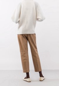 Maglione bianco a maglia con maniche a sbuffo, abbinato a pantaloni in pelle color cammello e ballerine beige, visto da dietro su uno sfondo chiaro.