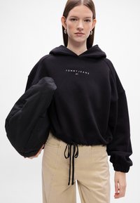 Felpa nera con logo bianco "TOMMY JEANS", orlo con coulisse e polsini a coste, abbinata a pantaloni beige e a una borsa nera.