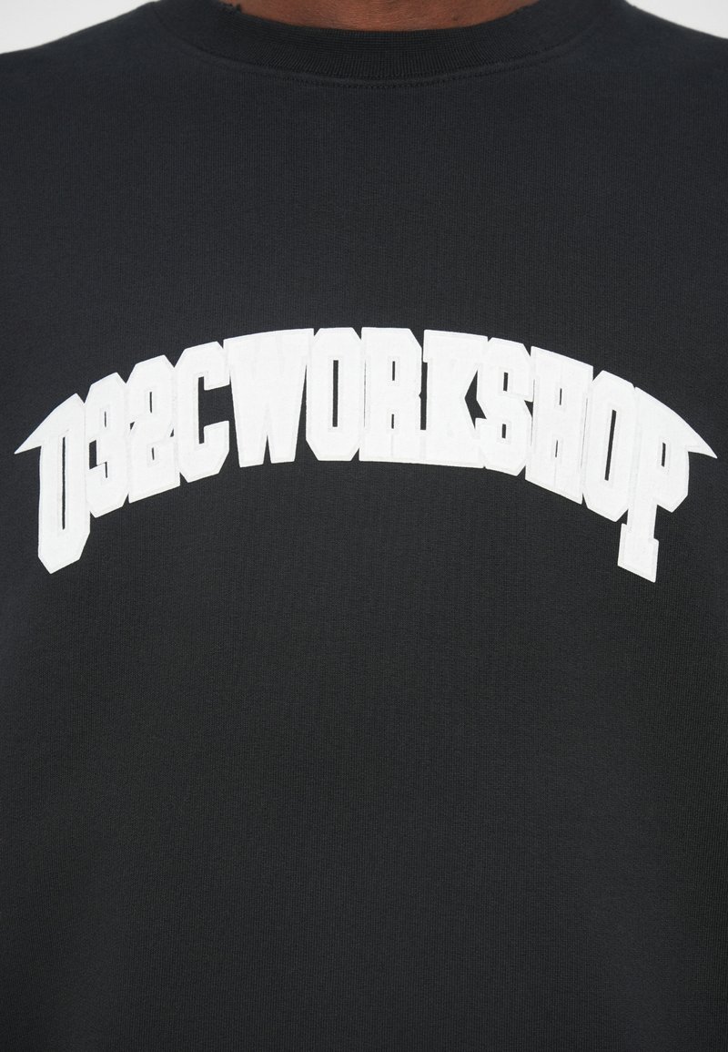 Zwarte shirt met witte dikke blokletters die "WORKSHOP" in een boogvorm over de borst spellen.