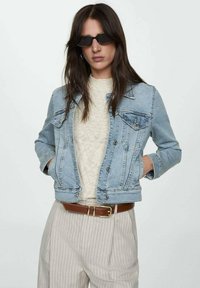 Lyseblå denimjakke med lange ærmer, brystlomme og sølvknapper; båret over en cremefarvet sweater og grå stribede bukser. Sorte solbriller.