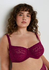 Donna dai capelli ricci che indossa un reggiseno di pizzo rosa scuro con spalline regolabili su uno sfondo chiaro e uniforme.