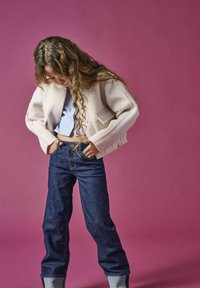 Giacca cropped rosa chiaro con due tasche frontali, abbinata a una camicia a righe blu e jeans in denim scuro con risvolti. Sfondo rosa.