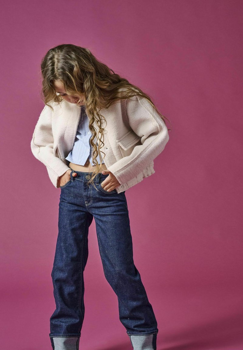 Giacca cropped rosa chiaro con due tasche frontali, abbinata a una camicia a righe blu e jeans in denim scuro con risvolti. Sfondo rosa.