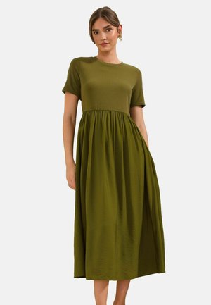 Femeie purtând o rochie midi verde măsliniu cu mâneci scurte, talie adunată și fustă fluidă, stând pe un fundal simplu și luminos.