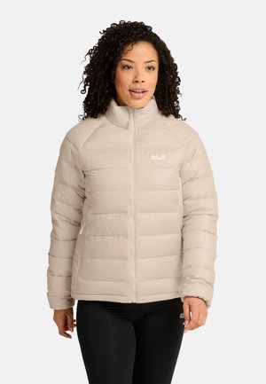Vrouw met krullend haar die een beige donsjas met hoge kraag en zwarte legging draagt tegen een effen achtergrond.