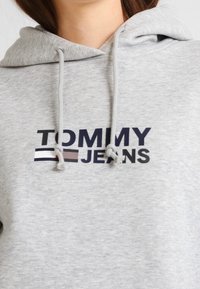 Felpa grigia con cappuccio e stringhe, caratterizzata da un grande logo "TOMMY JEANS" in blu scuro e rosso. Tessuto morbido e strutturato con una vestibilità comoda.