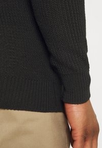 Svart stickad tröja med ett strukturerat mönster, med ribbade manschetter och nederkant, parat med ljusbeige tyg synligt nedanför.