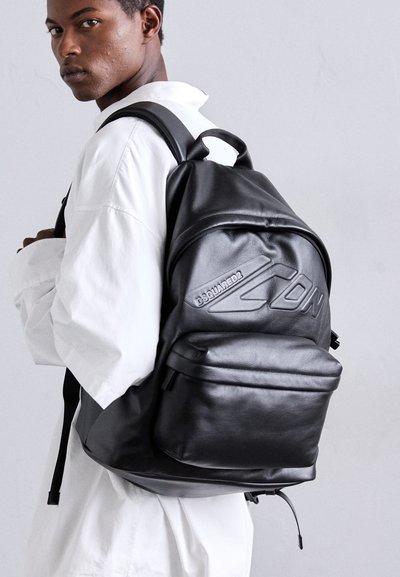 Black leather backpack with adjustable straps, distinct embossed logo, and front zip pocket; smooth texture and structured design.