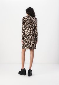Femme aux longs cheveux bouclés portant une robe mi-longue à motif léopard et des chaussures compensées noires, debout de dos sur un fond blanc.