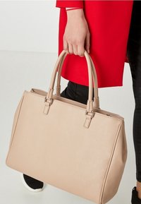 Borsa tote in pelle beige con due manici superiori, forma strutturata, dettagli di cucitura e chiusura con zip. Tenuta in una mano accanto a un cappotto rosso.