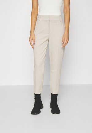 Femme portant un pantalon beige sur mesure longueur cheville, un haut blanc sans manches, et des bottines à talons noires sur un fond uni.