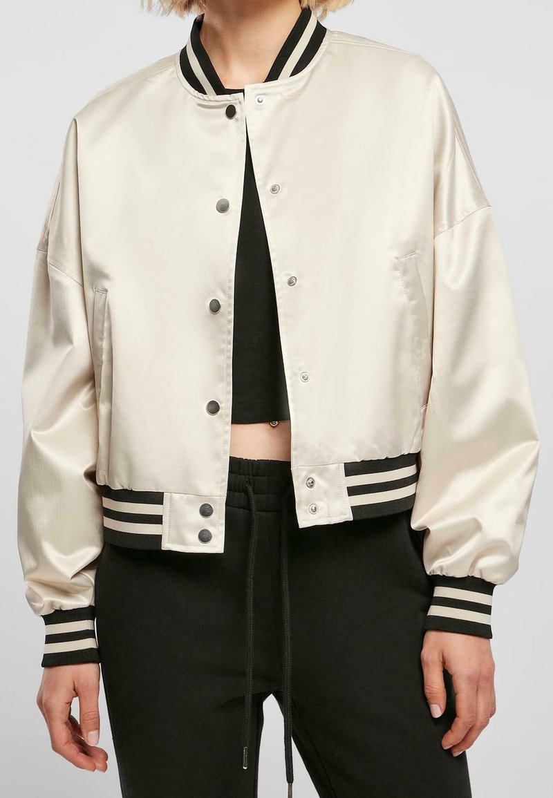 Veste bomber en satin coupée en crème avec bord côtes rayé noir, boutons-pression et poignets élastiques, associée à un pantalon de survêtement noir.