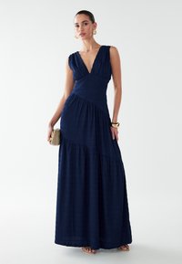 Marineblaues Maxikleid aus strukturiertem Stoff, mit tiefem V-Ausschnitt, tailliertem Oberteil und gestuftem Rock. Kombiniert mit goldenen Schmuckstücken und einer Clutch.