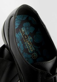 Schwarzer Lederschuh mit glatter Oberfläche, ausgestattet mit einer blauen, floral gemusterten Innensohle und goldener Schrift. Runde Form mit minimalistischem Design.
