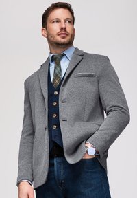 Homme portant un blazer gris, une chemise bleue, une cravate à carreaux, un gilet bleu marine, un jean et une montre-bracelet argentée regardant vers le haut avec une main dans la poche.