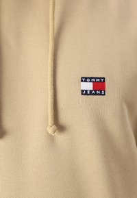 Beige hoodie with a textured finish, featuring a hood with drawstrings and a small navy and red "TOMMY JEANS" logo on the chest.