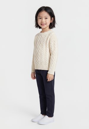 Παιδί με κοντά μαύρα μαλλιά φοράει μπεζ πουλόβερ με πλεκτό σχέδιο, σκούρο παντελόνι και λευκά παπούτσια χωρίς κορδόνια, στέκεται και χαμογελάει μπροστά σε απλό φόντο.