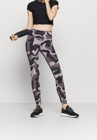 Svart avkortad topp, grå och svart kamouflagemönstrade leggings, och svarta träningsskor. Modellen håller en svart vattenflaska. Slät tyg, åtsittande design.