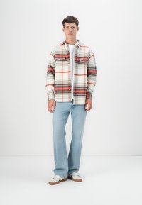 Camisa de cuadros en beige, gris y naranja; diseño de botón con dos bolsillos en el pecho. Combinada con jeans azul claro y zapatillas blancas.