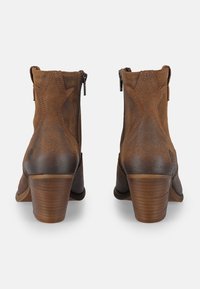 Botas de tobillo de cuero marrón con una textura suave, punta afilada y tacón curvado. Presentan costuras en contraste y un cierre de cremallera.