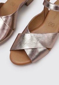 Sandalias metálicas de bronce con un diseño de correas cruzadas, material de cuero, con punta abierta y un tacón bajo y grueso. Textura suave con un brillo sutil.