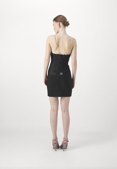 Robe mini noire avec des bretelles asymétriques, texture froncée sur un côté, et fermeture éclair dans le dos. Accompagnée de talons hauts avec des accents. 