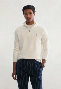 White zip-up sweater with knit texture, featuring a small logo on the chest, paired with navy pinstriped trousers.