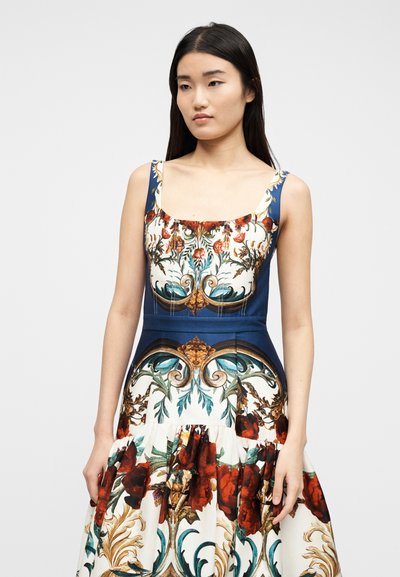 Femme aux longs cheveux foncés portant une robe sans manches à fleurs avec des motifs ornés rouges, bleus et dorés sur fond blanc, debout devant un arrière-plan uni.