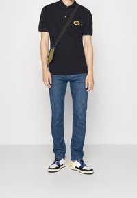 Černé polo tričko se žlutým logem, modré džíny a barevné tenisky. Oblečení doplňuje malá olivově zelená taška přes rameno.