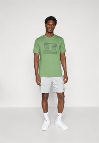 Grünes Baumwoll-T-Shirt mit einem schwarzen Grafik-Logo, kombiniert mit grauen Shorts. Das Modell trägt weiße Sneaker und Socken. Einfaches, lässiges Design.