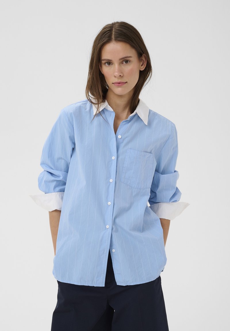 Žena s hnedými vlasmi, oblečená v svetlomodrej pruhovanej košeli s bielym golierom a manžetami, ruky v vreckách, na jednofarebnom pozadí.