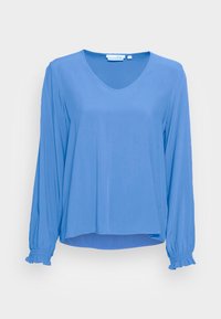 Blå långärmad blus med V-ringning, elastiska volangärmar och en något längre bakre fåll mot en enfärgad ljus bakgrund.