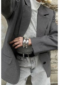 Personne portant un blazer gris, un col roulé clair, un jean clair, une ceinture noire et de gros bracelets argentés, debout près d’un mur texturé.