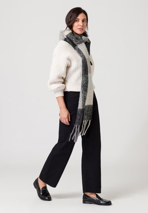 Donna che cammina indossando un maglione lavorato a maglia bianco, pantaloni neri, mocassini neri e una lunga sciarpa a quadri grigi e bianchi.