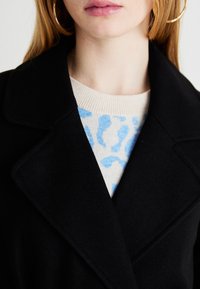 Μαύρο μάλλινο παλτό με κολάρο πέτο, φορεμένο πάνω από ένα ανοιχτό μπεζ πουλόβερ με μπλε αφηρημένο σχέδιο. Κοντινό στο λαιμό.