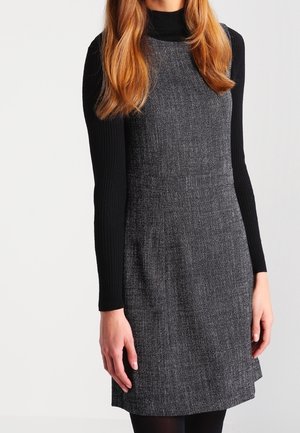 Frau trägt ein ärmelloses graues strukturiertes Kleid über einem schwarzen gerippten langärmligen Rollkragenpullover und steht vor einem schlichten hellen Hintergrund.