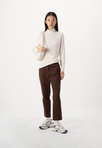 Lys beige høyhalset genser, brune rette bukser og metalliske joggesko. Modellen holder en veske med et mønstret stropp.