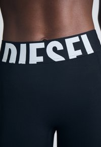 Des leggings noirs dotés d'une large taille élastique avec l'inscription blanche "DIESEL". Le tissu semble lisse et ajusté, mettant en valeur un design épuré.