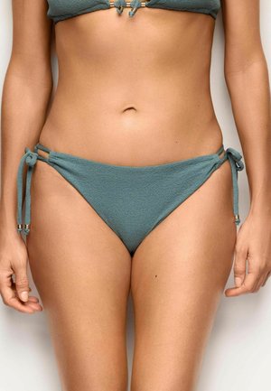 Donna che indossa slip bikini turchese testurizzato con legacci laterali e top bikini abbinato, in piedi davanti a uno sfondo chiaro e uniforme.