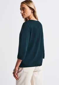 Blouse turquoise à manches longues avec une coupe décontractée, dotée d'un col rond et de détails froncés au niveau des poignets, associée à un pantalon crème.