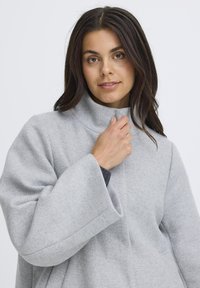 Grå uldblandingsfrakke med høj krave, brede ærmer og glat tekstur. Modellen holder i kraven og fremviser tøjets design.