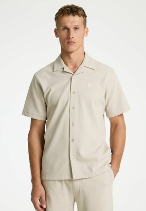 Jeune homme portant une chemise beige à manches courtes boutonnée et un pantalon assorti, debout avec une main dans la poche sur un fond uni.