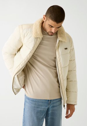 Puff-Jacke in Creme mit einem strukturierten, beigen Kunstfellkragen, ausgestattet mit einem Frontreißverschluss und horizontalem Steppmuster, getragen über einem beigen Shirt.