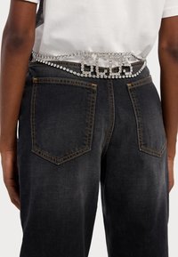 Strieborný opasok s kamienkami má veľké prepojené logo s dekoratívnym dizajnom reťaze a nosí sa cez tmavé denimové nohavice s kontrastným prešívaním.